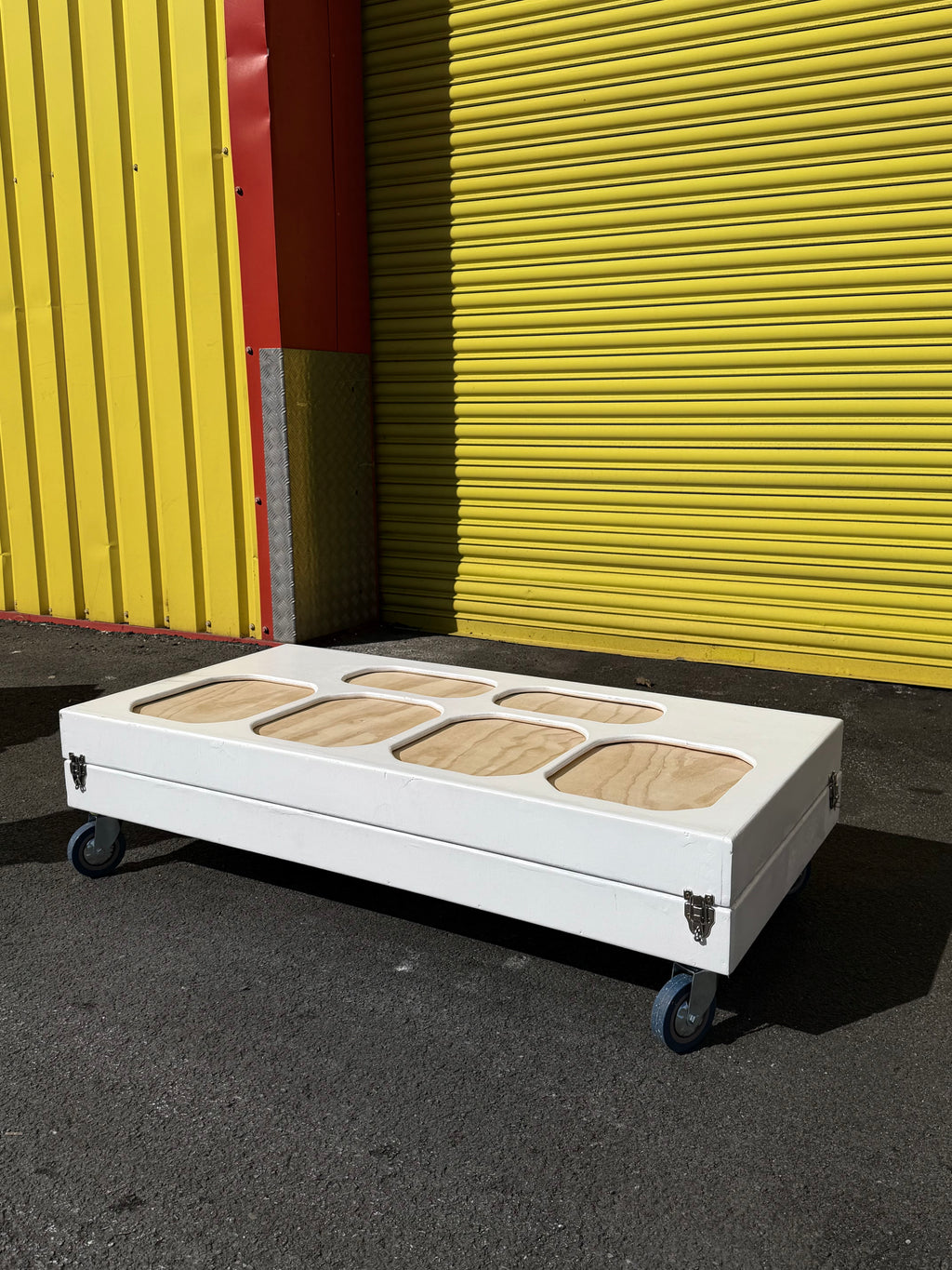 A white rectangular rolling storage box with six oval cutouts on top, showing light wood inserts, set on a black wheeled dolly in front of a bright yellow corrugated metal wall.