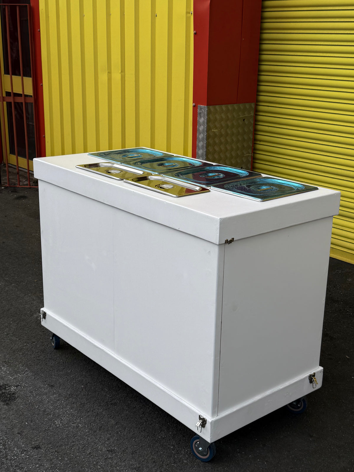 A white commercial ice cream freezer cart with four stainless steel compartments on top, mounted on blue caster wheels, photographed outdoors against a yellow corrugated metal wall.